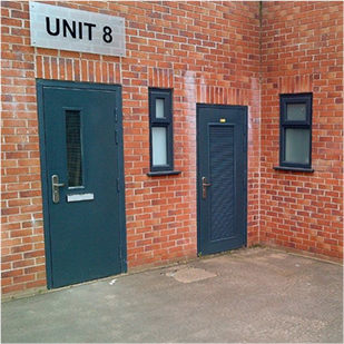 Two grey doors on a brick building one with a full louvre the other a small window under a grey sign saying unit 8 in black